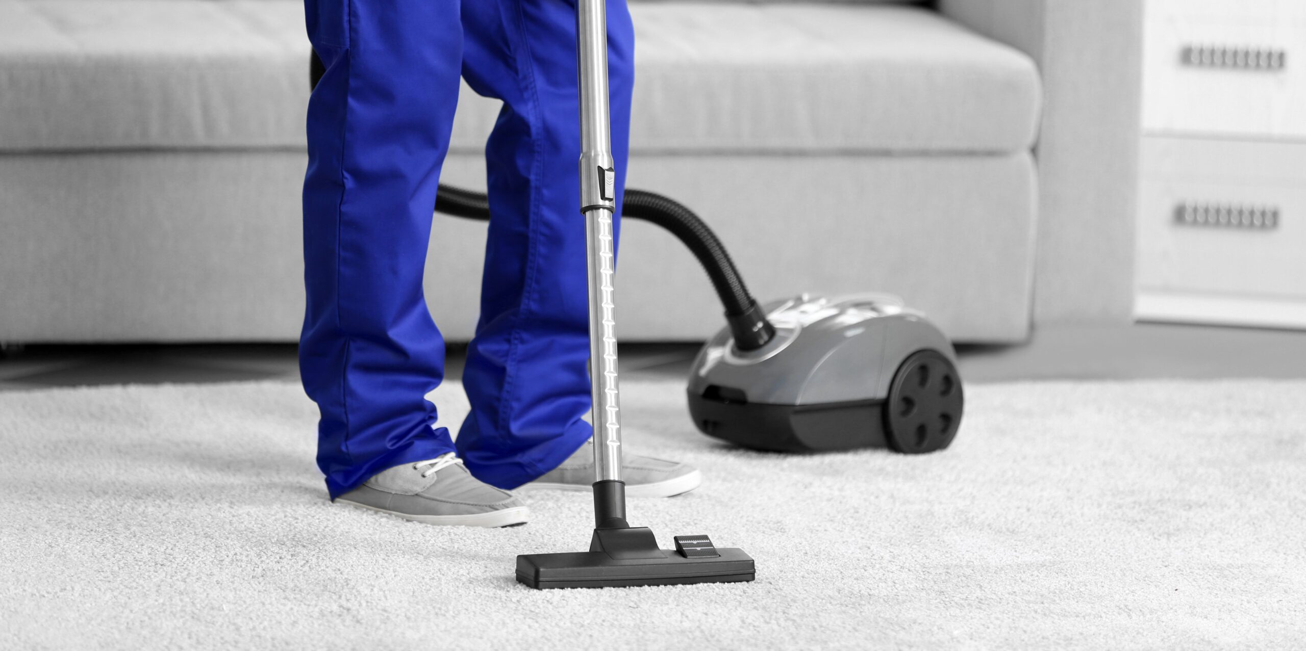 A person wearing blue pants and a blue shirt is cleaning a carpet with a vacuum cleaner