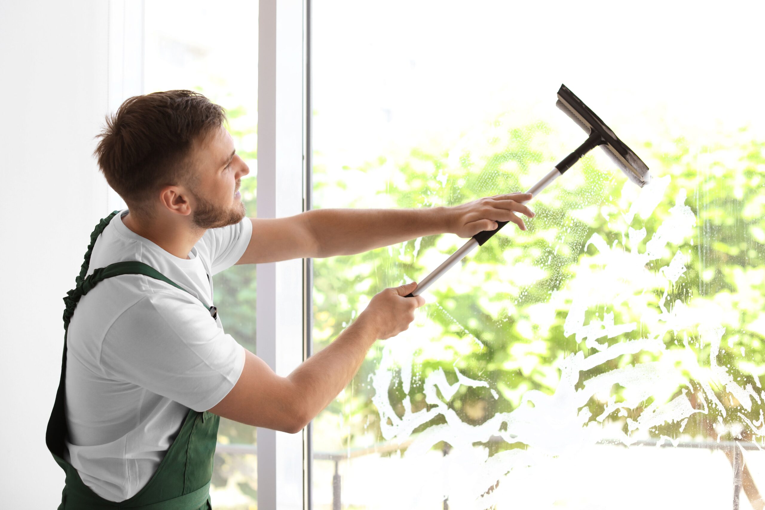 A man in overalls uses a mop to clean a large window, focusing on removing dirt and streaks