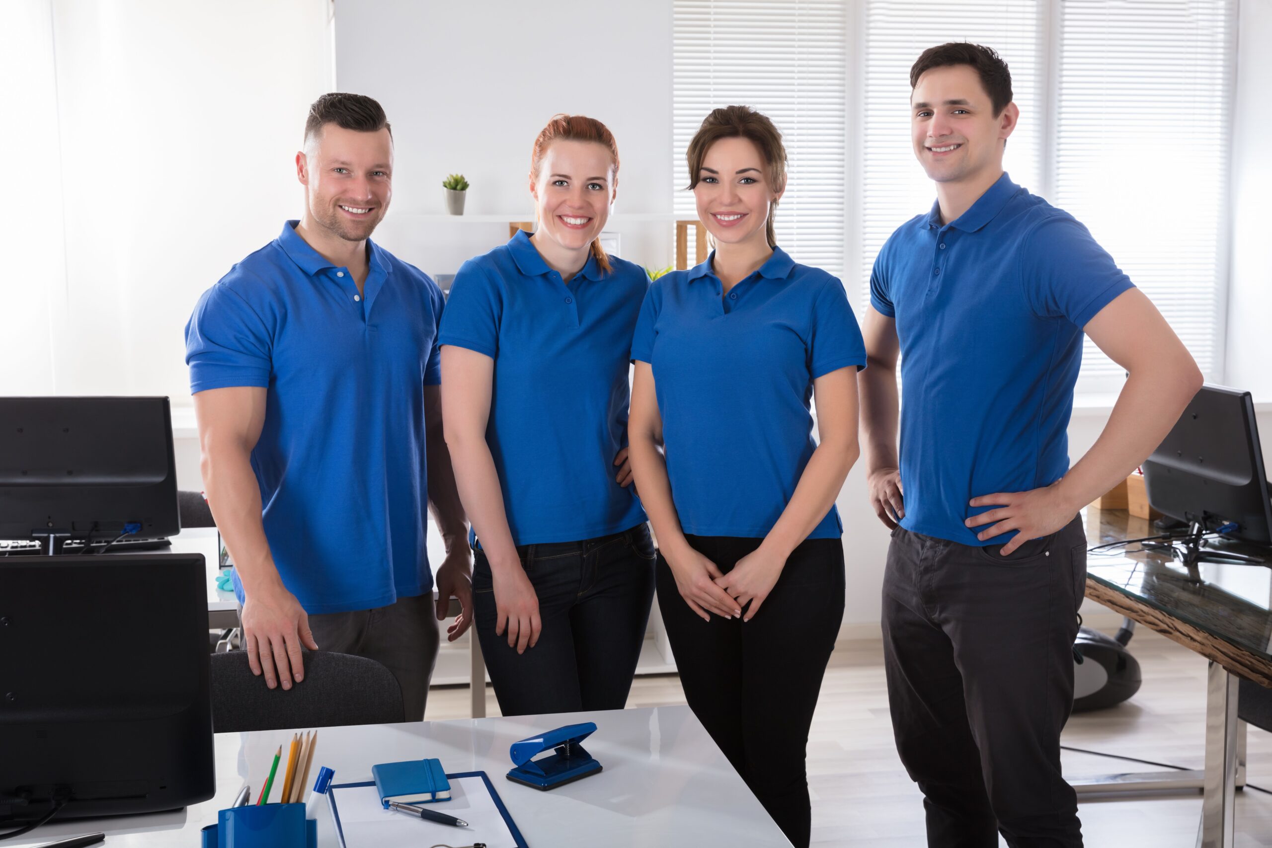 four individuals in blue shirts stand in front of a desk, engaged in conversation or collaboration