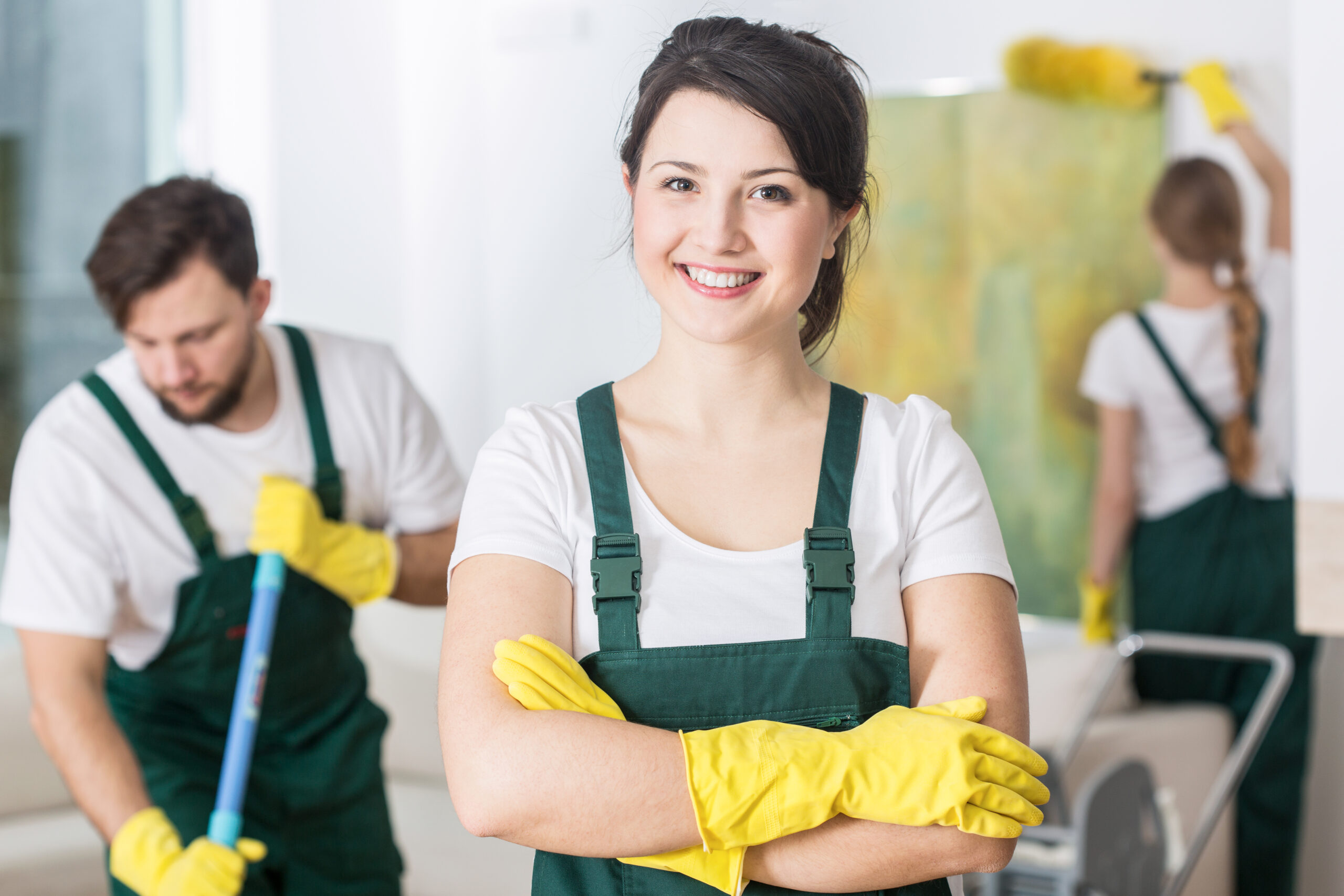 A woman in overalls and yellow gloves stands before a group of people, engaging them with a friendly demeanor